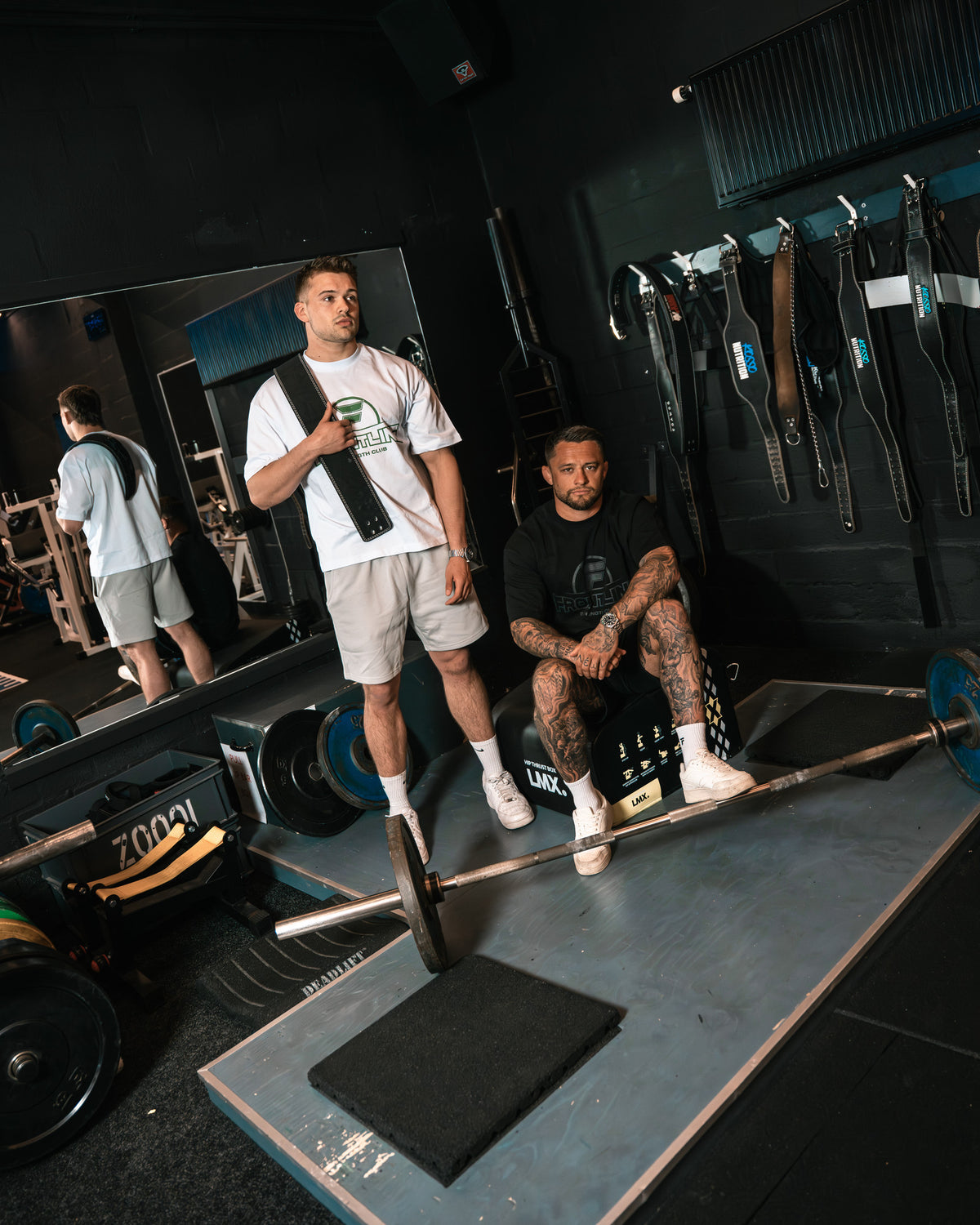 Strengthclub T-Shirt wit met een lifting belt over de schouder, naast het zwarte shirt